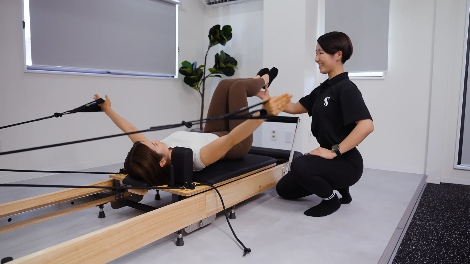 リフォーマーで仰向けになり腕を動かす利用者と女性トレーナー